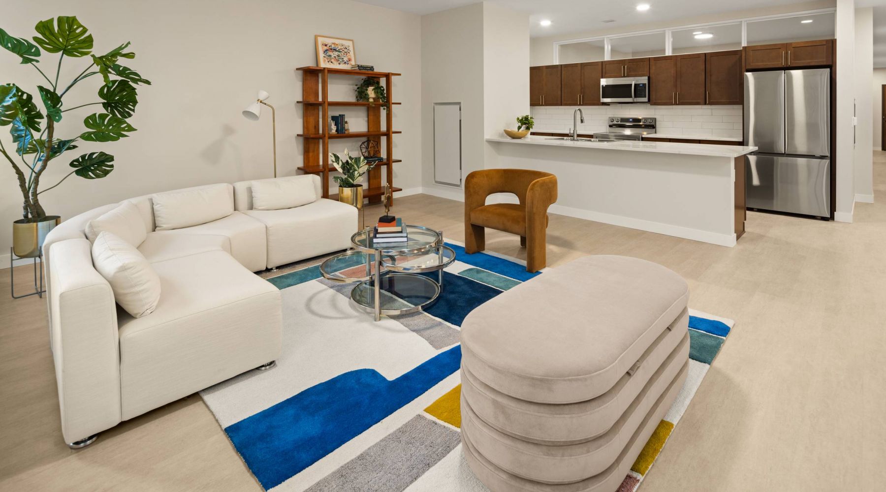 Modern living area in Groove Lofts featuring a cozy white sectional sofa, glass coffee table, colorful geometric rug, and a view of the open kitchen with stainless steel appliances and dark wood cabinets.